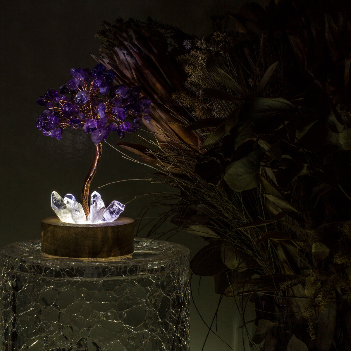 Indulge in a peaceful night&#39;s sleep next to a stunning crystal tree of life, carefully crafted to promote energetic healing and a serene ambiance. Immerse yourself in the gentle, calming glow emanating from the precisely cut clear quartz points, arranged meticulously on a natural, visually appealing wooden base. Accentuating the entire structure is a delicate copper wire stem, elegantly adorned with your choice of crystal chip leaf topping.