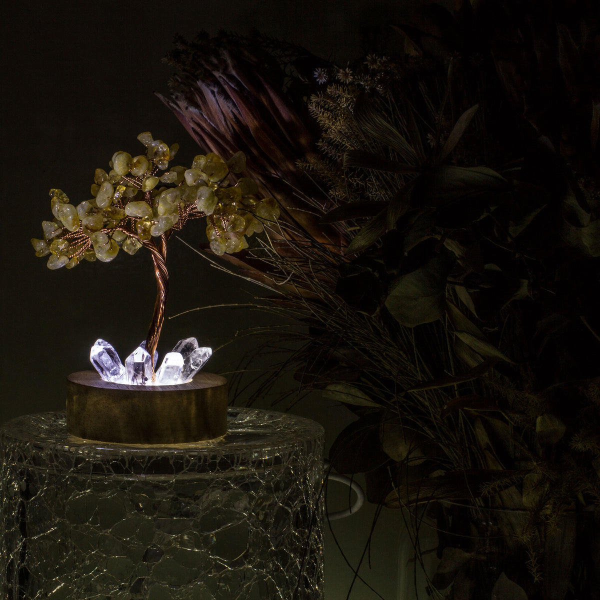 Indulge in a peaceful night&#39;s sleep next to a stunning crystal tree of life, carefully crafted to promote energetic healing and a serene ambiance. Immerse yourself in the gentle, calming glow emanating from the precisely cut clear quartz points, arranged meticulously on a natural, visually appealing wooden base. Accentuating the entire structure is a delicate copper wire stem, elegantly adorned with your choice of crystal chip leaf topping.