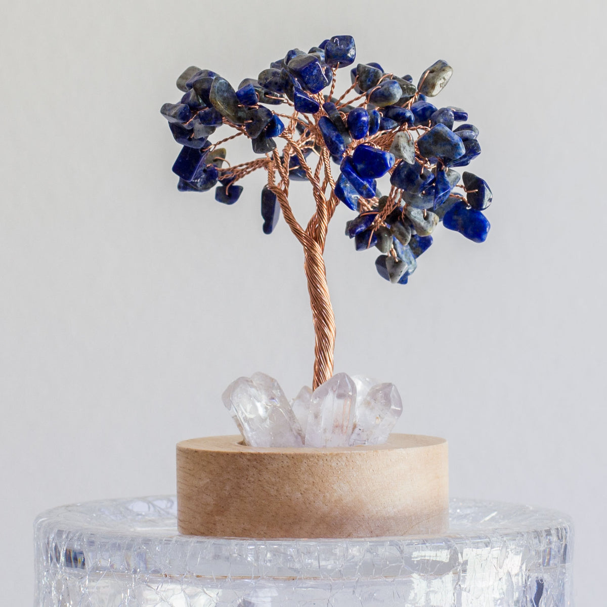 Indulge in a peaceful night&#39;s sleep next to a stunning crystal tree of life, carefully crafted to promote energetic healing and a serene ambiance. Immerse yourself in the gentle, calming glow emanating from the precisely cut clear quartz points, arranged meticulously on a natural, visually appealing wooden base. Accentuating the entire structure is a delicate copper wire stem, elegantly adorned with your choice of crystal chip leaf topping.