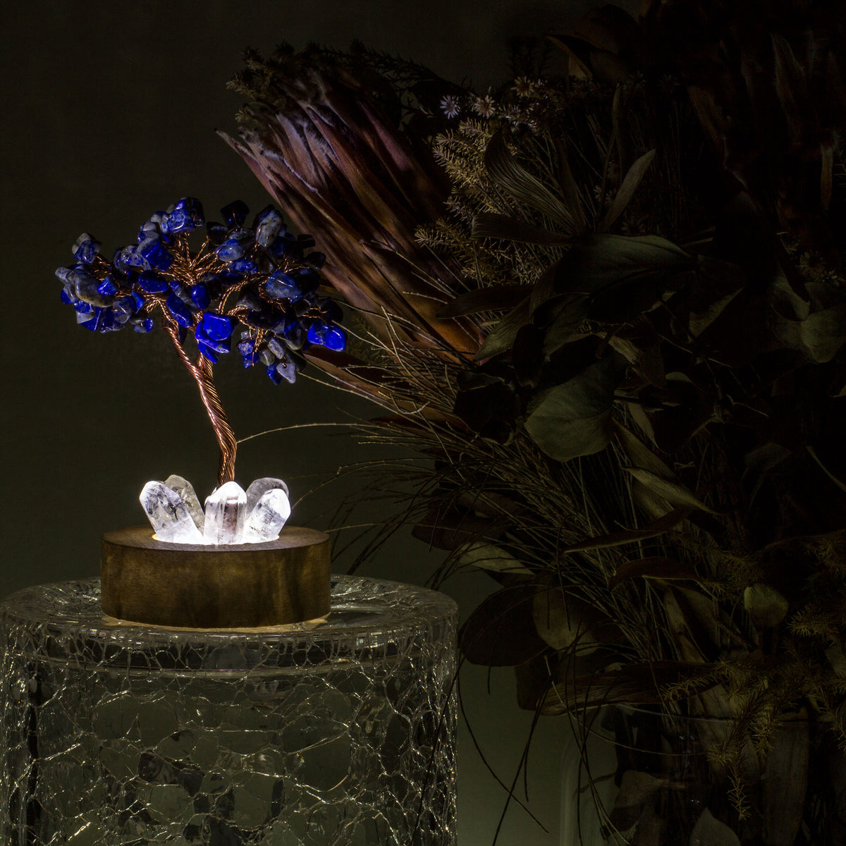 Indulge in a peaceful night&#39;s sleep next to a stunning crystal tree of life, carefully crafted to promote energetic healing and a serene ambiance. Immerse yourself in the gentle, calming glow emanating from the precisely cut clear quartz points, arranged meticulously on a natural, visually appealing wooden base. Accentuating the entire structure is a delicate copper wire stem, elegantly adorned with your choice of crystal chip leaf topping.