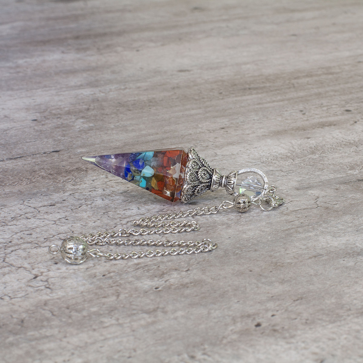 Pendulum - 7 Ckakra - Resin Mi Chakra A crystal pendulum is a simple tool commonly used for divination and dowsing. It typically consists of a crystal or gemstone suspended on a chain or cord. The crystal is usually pointed, allowing it to swing freely and respond to questions. Pendulums are believed to connect with your subconscious mind and energy fields, offering insights through their movements.- 