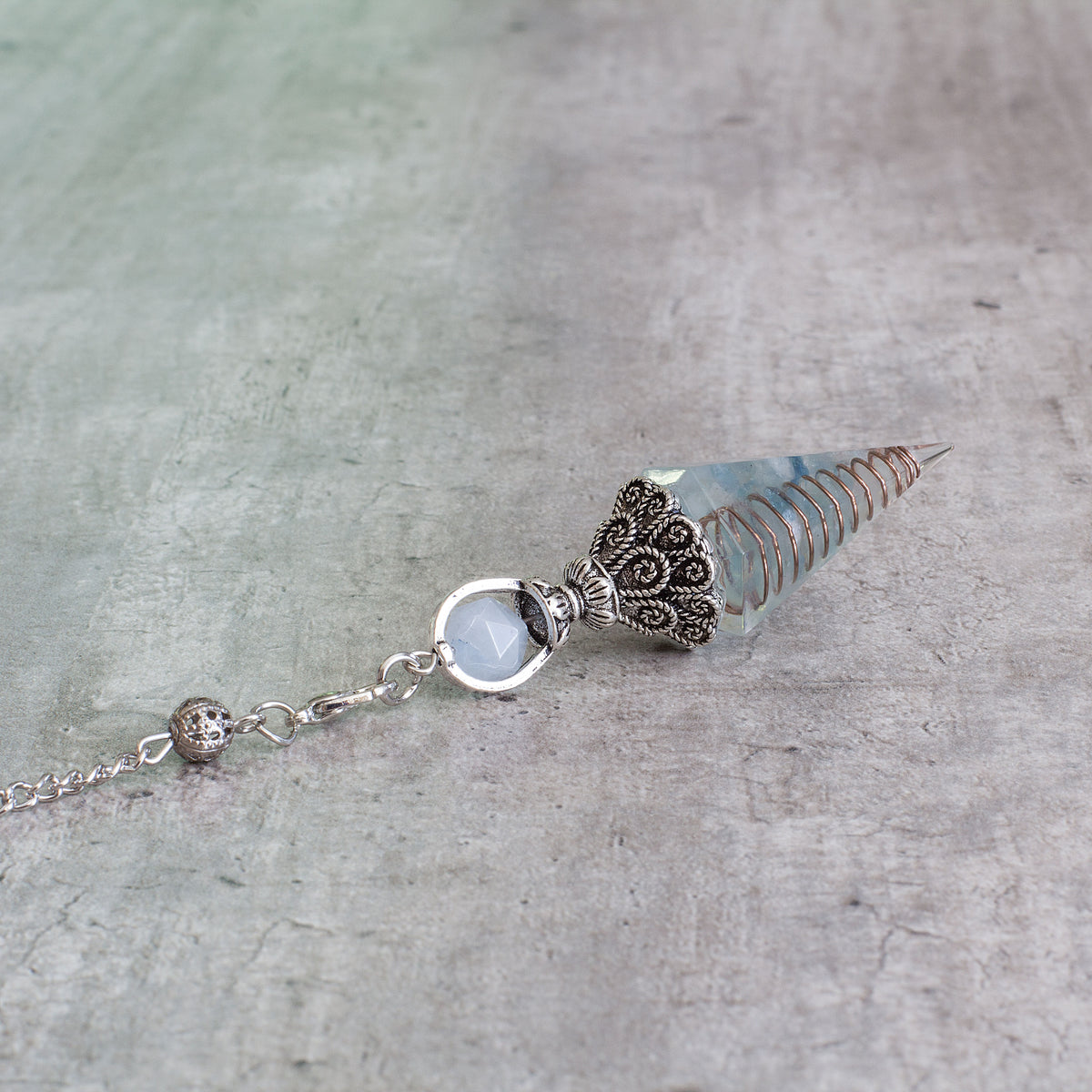 Pendulum - Aquamarine - Resin Mi Chakra - A crystal pendulum is a simple tool commonly used for divination and dowsing. It typically consists of a crystal or gemstone suspended on a chain or cord. The crystal is usually pointed, allowing it to swing freely and respond to questions. Pendulums are believed to connect with your subconscious mind and energy fields, offering insights through their movements.
