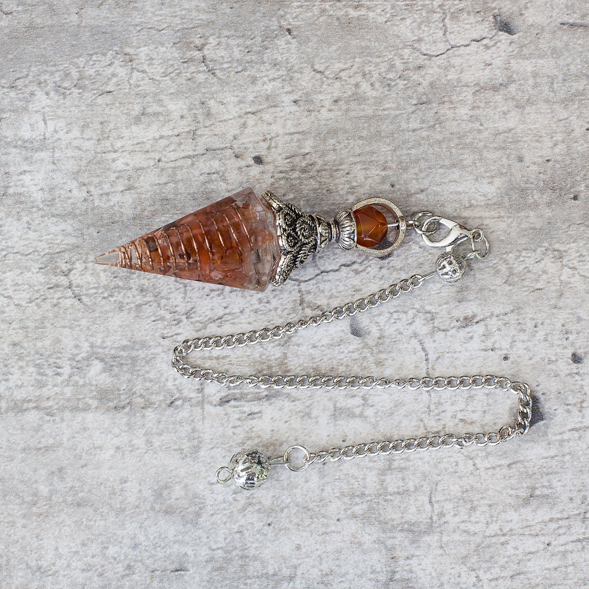 Pendulum - Carnelian - Resin Mi Chakra - A crystal pendulum is a simple tool commonly used for divination and dowsing. It typically consists of a crystal or gemstone suspended on a chain or cord. The crystal is usually pointed, allowing it to swing freely and respond to questions. Pendulums are believed to connect with your subconscious mind and energy fields, offering insights through their movements.