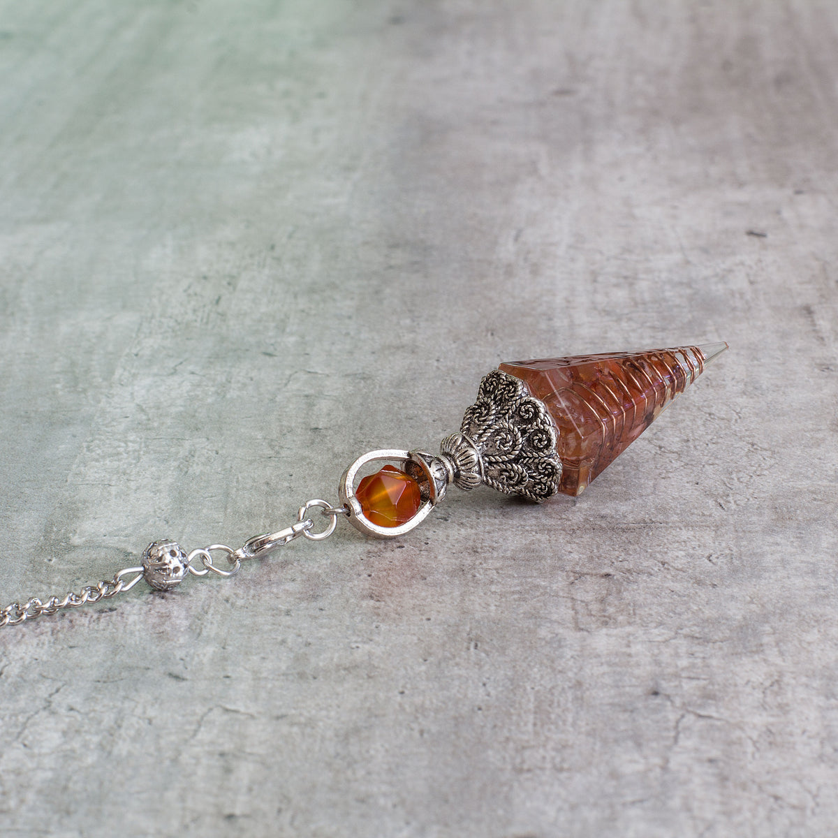 Pendulum - Carnelian - Resin Mi Chakra - A crystal pendulum is a simple tool commonly used for divination and dowsing. It typically consists of a crystal or gemstone suspended on a chain or cord. The crystal is usually pointed, allowing it to swing freely and respond to questions. Pendulums are believed to connect with your subconscious mind and energy fields, offering insights through their movements.