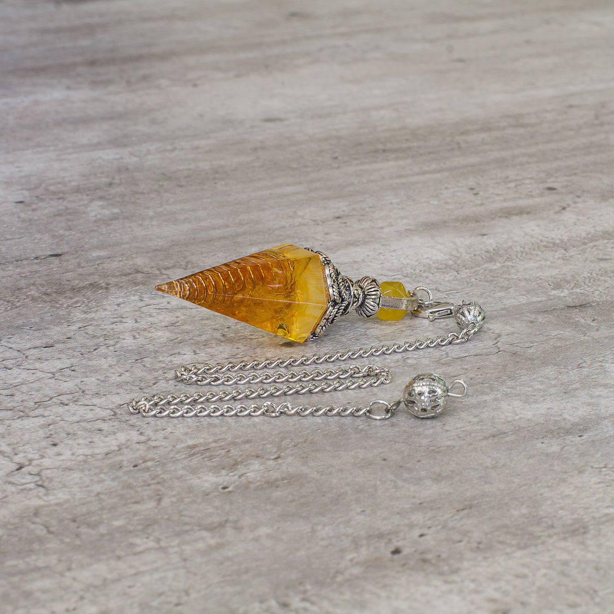 Pendulum - Citrine - Resin Mi Chakra - A crystal pendulum is a simple tool commonly used for divination and dowsing. It typically consists of a crystal or gemstone suspended on a chain or cord. The crystal is usually pointed, allowing it to swing freely and respond to questions. Pendulums are believed to connect with your subconscious mind and energy fields, offering insights through their movements.