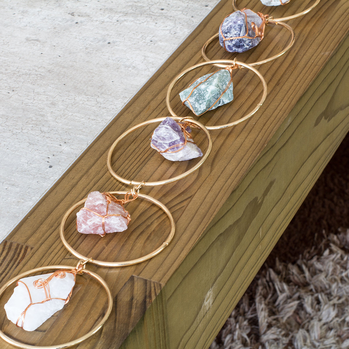 Turn your home into a haven of beauty and tranquility with this exceptional wall art meticulously handcrafted from genuine rough natural stones. Seven stunning, hand-selected chunks of Rose Quartz, Quartz Crystal, Green Aventurine, Amethyst, Fluorite, Amazonite, and Strawberry Quartz are carefully wrapped and suspended from an elegant, intricately detailed golden iron ring, creating a truly one-of-a-kind statement piece that will add an air of sophistication and natural charm to any room in your home.